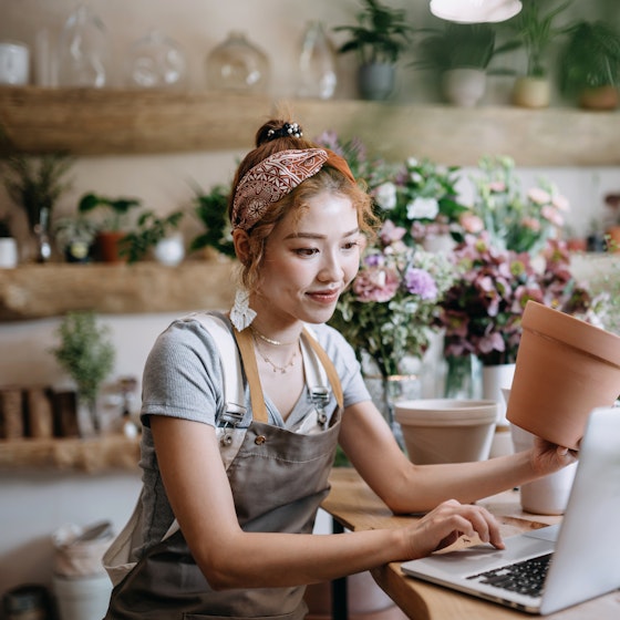 A young woman sits at a wood-topped counter in a plant shop and looks at something on the screen of an open laptop. With one hand, the woman holds a clay pot; with the other, she manipulates the track pad on the laptop. The woman wears a gray apron over a light blue T-shirt. Her hair is pulled up in a bun and partially covered by a paisley-patterned headband. Most of the counter is covered with potted plants and flowers, as are the wooden shelves on the wall behind the woman.