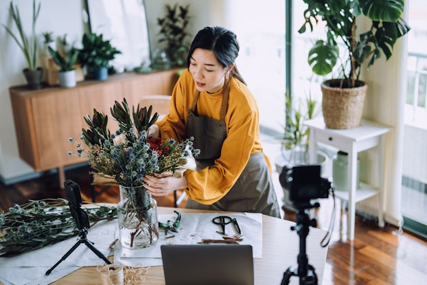 A woman adjusts a flower arrangement in a glass vase on a table. In the foreground, out of focus, a smartphone on a tripod is pointed at the woman; another smartphone on a shorter tripod sits on the table, pointed directly at the vase. The woman wears a mustard yellow sweater under a gray apron. The flower arrangement is made up succulents with long dark green leaves, blue plants with small fuzzy buds, and a cluster of small, bright red flowers. Extra plants lay on the table, along with a couple of pairs of pruning shears and some cut-off stems. In the background, various potted plants stand on a side table and a dresser.