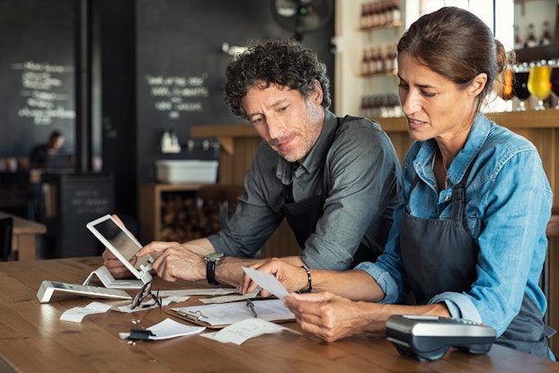  A couple sits side by side at a wooden table scattered with papers, receipts, and a couple of calculators. The man and woman wear matching dark blue aprons and look worried. The woman sitting on the right looks down at a receipt in her hand. The man on the left also looks at the receipt as he taps at an electronic tablet. In the background is a wine bar. A wooden bar holding several colored wine glasses stands behind the sitting couple; behind the bar are shelves of wine bottles. Another wall, running to the left of the couple, is a chalkboard wall with writing on it.