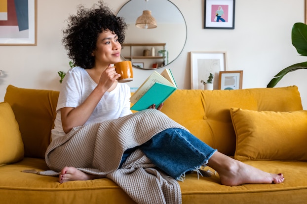 A curly-haired woman sits on a mustard yellow couch with a book in one hand and a mug in the other. She is barefoot and has a blanket draped over her lap. She looks off-screen with a content expression.