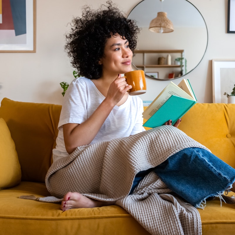 A curly-haired woman sits on a mustard yellow couch with a book in one hand and a mug in the other. She is barefoot and has a blanket draped over her lap. She looks off-screen with a content expression.