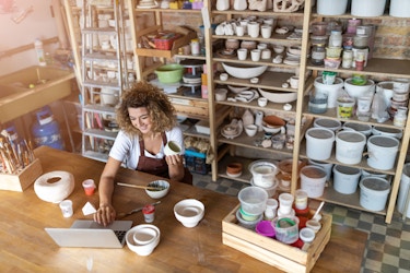  A woman works on her laptop at a large wooden table scattered with pottery. Behind her are shelves of handmade bowls, pots, buckets, and other pieces of pottery. 