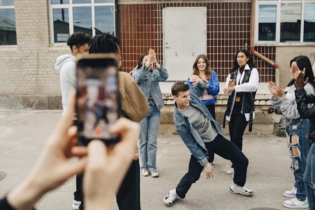 A group of teens stands in a circle outside, applauding one teen in the center of the circle. The teen in the center is a boy in black pants, a gray shirt, a denim jacket, and black-and-white sneakers. The boy is mid-dance, bending himself backward with his legs slightly bent and one arm extending upward. In the foreground, out of focus, a pair of hands holds up a smartphone and films the scene. Behind the group of teens is a building with a tan brick exterior and concrete steps leading up to a white door.