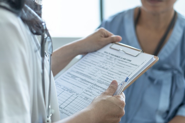 A shot from the neck down showing a person holding a form on a clipboard and a pen and another person wearing medical scrubs.