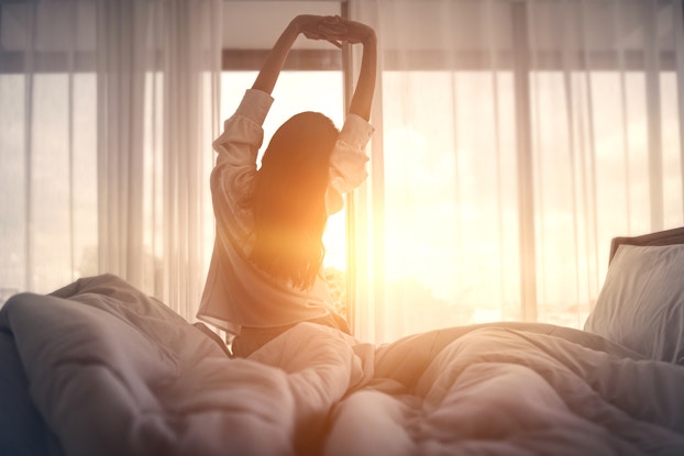 Person stretching in bed with the sun peeking through the curtains.