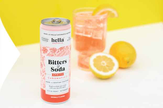 Can of Hella Cocktail Co. bitters & soda on a table with a full glass and a lemon,