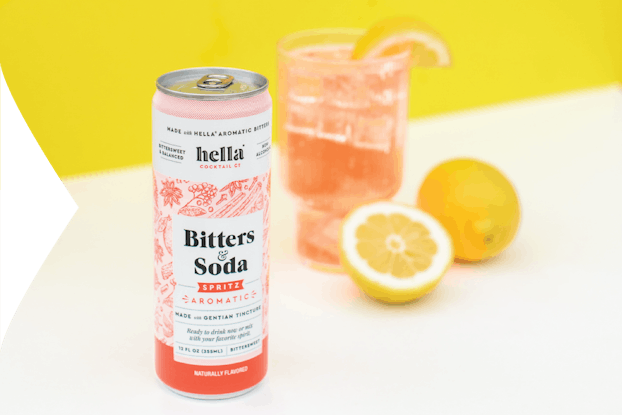  Can of Hella Cocktail Co. bitters & soda on a table with a full glass and a lemon,