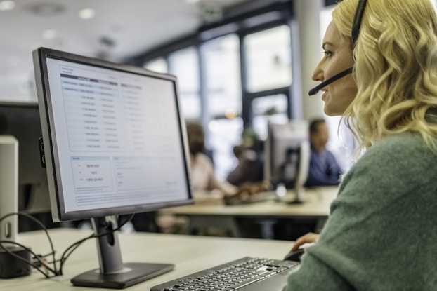 A profile shot of blonde woman wearing a headset with a microphone extension. She sits at a desk and looks at a computer monitor, which shows a list with three columns. The woman has one hand on a computer mouse on the desk and is speaking into the headset. In the background, other people sit at similar computer setups at other desks.