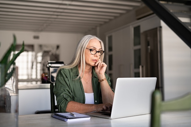  A woman in a minimalist room sits at a table and looks at a laptop. One hand is on her chin and she looks thoughtful.