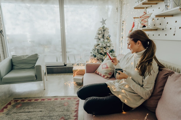 Woman sitting on her couch on her phone in an apartment decorated for Christmas.