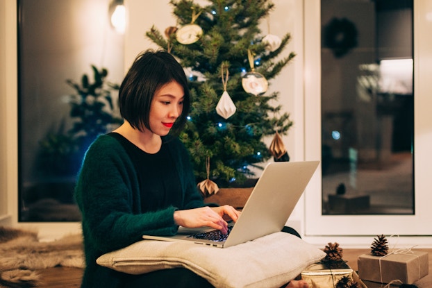 A woman sits on the floor in front of a Christmas tree. On her lap is a laptop on top of a cream-colored pillow. The woman wears a dark green sweater and types on the laptop. On the floor around the woman and the Christmas tree are a few boxes wrapped in silver and gray paper, tied up with string, and decorated with pinecones.