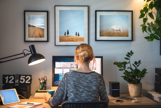  A woman seen from behind works at a home office. Her upper body partially obscures a computer screen and her desk is scattered with various electronics, office supplies and potted plants. Three large framed photographs on the wall behind the desk show scenes of prairie fields under open skies.