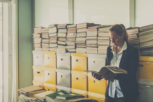  Business woman stands in an office room full of documents in boxes and stacked on top of them.