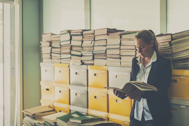 Business woman stands in an office room full of documents in boxes and stacked on top of them.