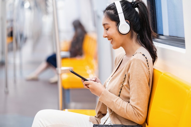 Woman on a train wearing headphones and smiling down at her phone.