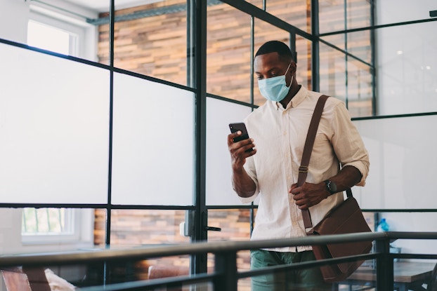  Man walking through office wearing a mask and looking at his phone.