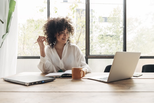  A woman sits at a wooden table with her back to a bank of windows. An open notebook and a coffee mug sit in front of the woman, who holds a pencil and looks at an open laptop with a smile.