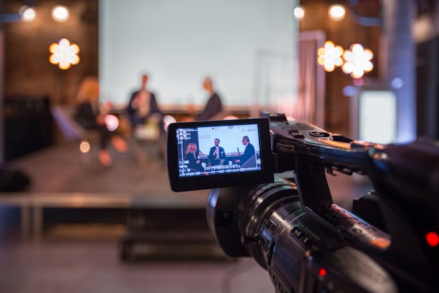  Video camera recording a live business event with people sitting on a stage.
