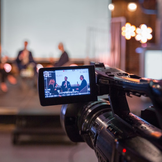  Video camera recording a live business event with people sitting on a stage. 