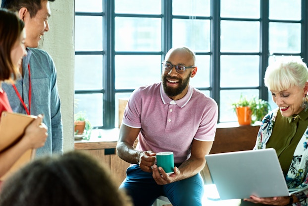  A group of coworkers stand and sit in an open area next to a bank of windows. Three people are standing at the edge of the image, facing away from the viewer and toward the person in the center of the image. The person in the center of the image is a smiling bald man with a beard; he is wearing a lavender polo shirt and glasses and is holding a teal coffee mug. The man is seated on the edge of a table. Next to him, also sitting on the edge of the table, is an older woman holding an open laptop. The woman has short white-blonde hair and is wearing a leaf-patterned jacket over a zip-up olive green shirt.