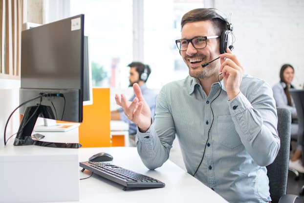 A man wearing a button-up shirt, glasses and a headset sits at a desk in front of a computer monitor and keyboard. He uses one hand to hold the mouthpiece of the headset and is laughing.