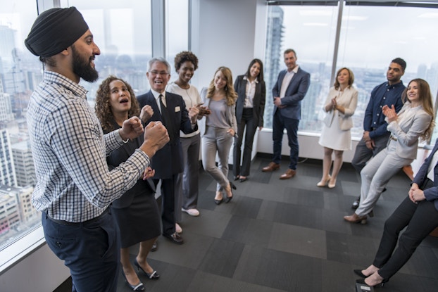  A group of about a dozen people in businesswear stand in a wide circle in an empty area of an office. The group's attention is on the man standing on the far left; he has a dark beard and wears a turbin and a windowpane plaid shirt. His fists are clenched in front of him in excitement and a couple of the other members of the group are applauding. The floor-to-ceiling windows behind the group show a sprawling cityscape.