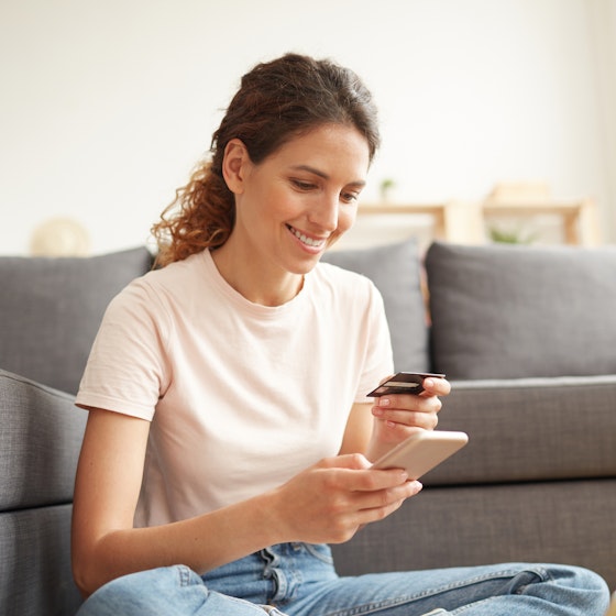 A young woman with her hair in a ponytail sits on the floor in front of a gray couch. She holds a smartphone in one hand and a credit card in the other, and looks down at both with a smile.