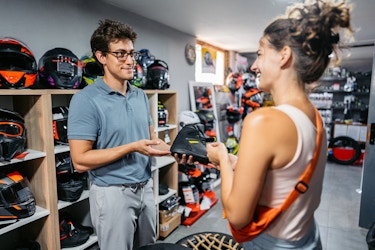 A man and a woman stand inside a sporting goods store. The man is an employee, wearing glasses, a teal polo shirt, and gray pants. He's holding a black athletic sneaker out to the woman. The woman is wearing a white tank top and has an orange bag slung over one shoulder. She is smiling at the employee. In the background are cubby-like shelves holding motorcycle helmets.