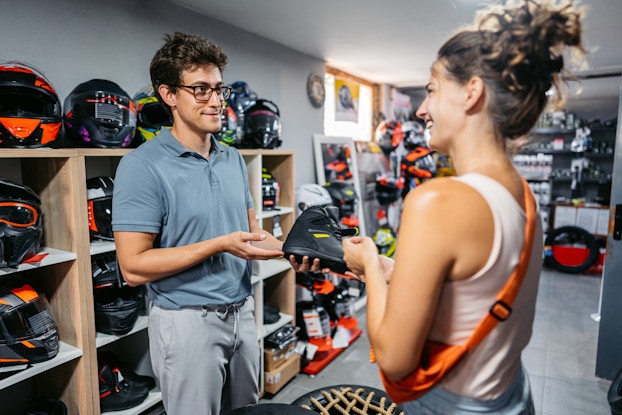 A man and a woman stand inside a sporting goods store. The man is an employee, wearing glasses, a teal polo shirt, and gray pants. He's holding a black athletic sneaker out to the woman. The woman is wearing a white tank top and has an orange bag slung over one shoulder. She is smiling at the employee. In the background are cubby-like shelves holding motorcycle helmets.