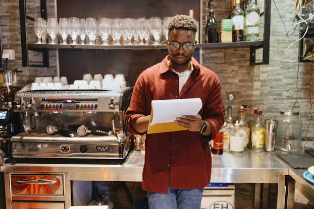 A coffee shop owner leans against a counter with a notebook in his hand. He is reviewing the inventory for his cafe.
