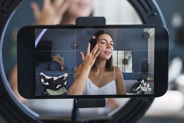  A close-up of a horizontal smartphone, the screen of which shows a woman rubbing lotion onto her cheek. The woman has freckled skin and dark, shoulder-length hair. She is seen from the chest up and is wearing a white camisole and a gold pendant. A pink hair clip holds her hair out of her face. The woman standing in a room with navy blue walls. The smartphone sits on a tripod in the middle of a ring light. In the background behind the filming phone is the woman seen on the screen, larger and out of focus.