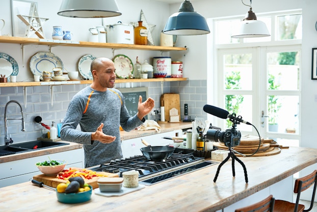  Man in his kitchen cooking food and recording himself on his phone.