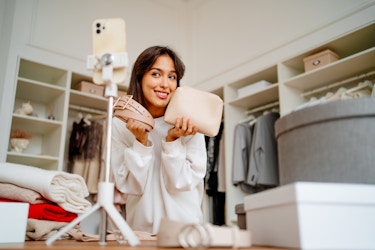  A smiling young woman stands across from a smartphone mounted on a tripod and poses with a pale pink belt and a cream-colored clutch held near her face. In the background are neutral walls into which shelves and alcoves are built. The alcolves are filled with clothes on hangers and the shelves hold pale pink storage boxes and mannequin heads displaying hats. 