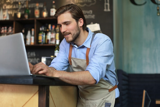 A man in a khaki apron and light blue button-up shirt leans on a counter at a cafe or bar and types something on a laptop. The man has brown hair and a brown beard. Behind him is a set of shelves filled with various bottles.