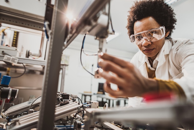 A woman wearing safety goggles and a white lab coat leans over an industrial machine. She uses one hand to adjust a piece of the machine that is connected with two wires.