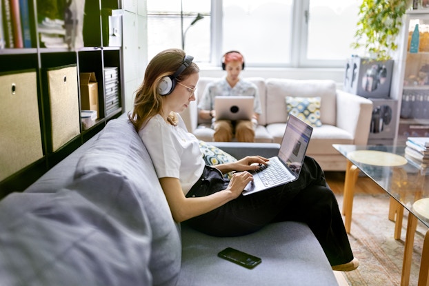 People working in an office on separate couches wearing headphones and on their laptops.