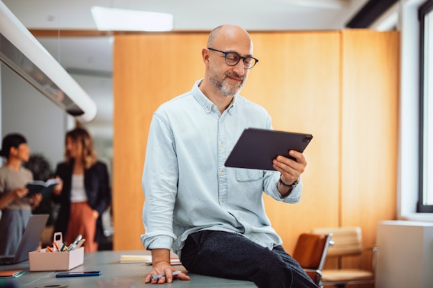 A male business owner leans against a desk in an office. He is reading an article on a computer tablet and smiling slightly.