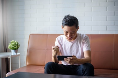  A young man wearing a white T-shirt sits on a brown leather couch and looks down at the smartphone in his hand. He smiles and pumps his fist in victory. 