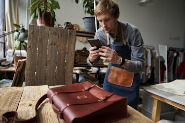  A man in a workshop uses a smartphone to take a picture of a leather bag. The bag is a messenger bag with two buckles on the front; it's sitting on a table made of wooden planks. The man has blond hair and is wearing a light blue button-up shirt under a dark blue apron with a leather pocket on front. Behind him in the workshop are racks of clothing and shelves holding a couple of potted plants. 