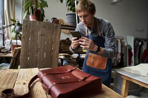  A man in a workshop uses a smartphone to take a picture of a leather bag. The bag is a messenger bag with two buckles on the front; it's sitting on a table made of wooden planks. The man has blond hair and is wearing a light blue button-up shirt under a dark blue apron with a leather pocket on front. Behind him in the workshop are racks of clothing and shelves holding a couple of potted plants.