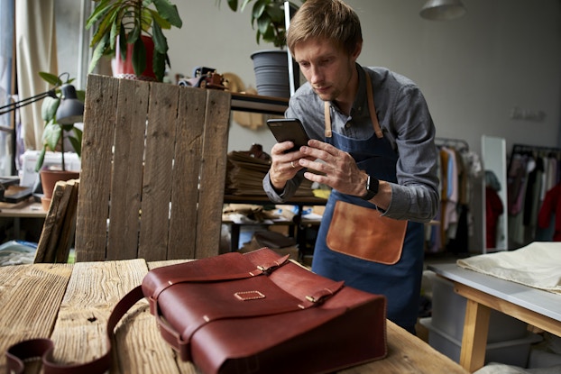 A man in a workshop uses a smartphone to take a picture of a leather bag. The bag is a messenger bag with two buckles on the front; it's sitting on a table made of wooden planks. The man has blond hair and is wearing a light blue button-up shirt under a dark blue apron with a leather pocket on front. Behind him in the workshop are racks of clothing and shelves holding a couple of potted plants.