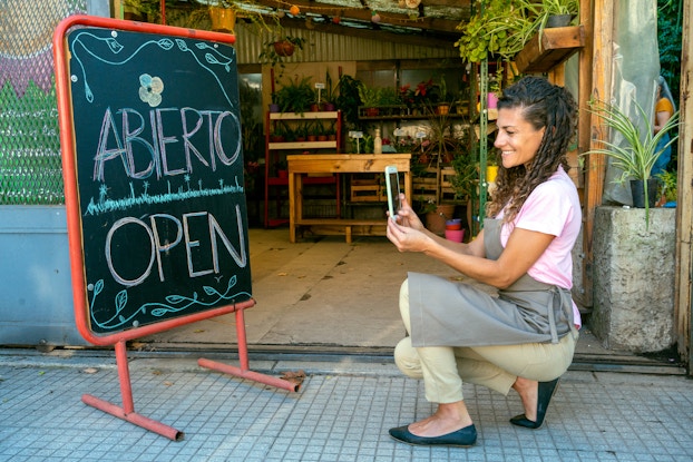 A woman kneels outside of a storefront, using a smartphone to take a picture of a chalkboard sign reading "ABIERTO | OPEN." The woman is wearing a gray apron over khakis and a light pink T-shirt. She has long curly hair and is smiling widely. The chalkboard sign has leafy vines drawn in green chalk around the text, and the I in "abierto" is dotted with a multicolored flower. The inside of the store is filled with potted plants, some hanging from the ceiling.