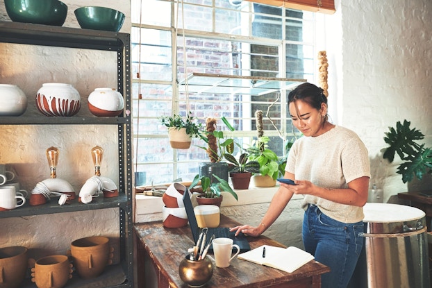 A woman stands next to a wooden table and looks at the smartphone in her hand. Her other hand rests on the keyboard of an open laptop on the table. On the other side of the table is a shelving unit filled with pottery. The table also holds some pots, including a small metallic gold pot filled with writing utensils. The windowsill next to the table is filled with potted plants.