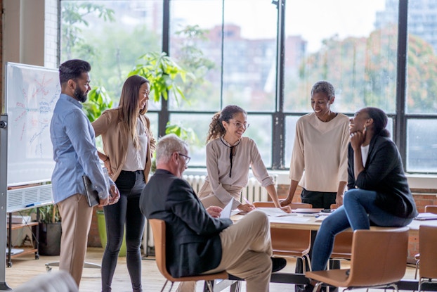 A group of employees are gathered around a small table while a woman speaks to the group. The image reflects a small, informal meeting of employees.