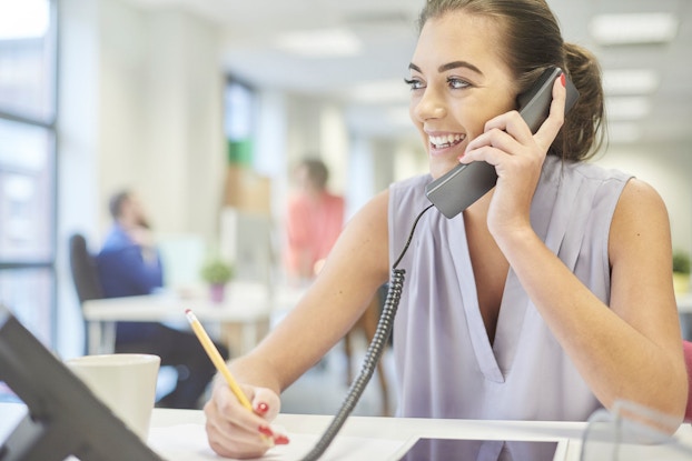 A young female intern pictured in a modern office is smiling and talking on the phone to a client.