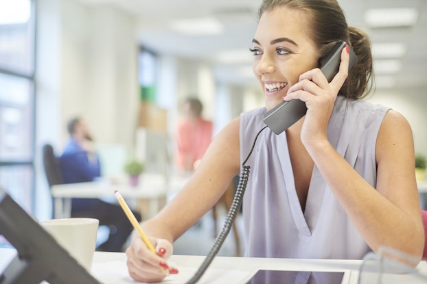  A young female intern pictured in a modern office is smiling and talking on the phone to a client.