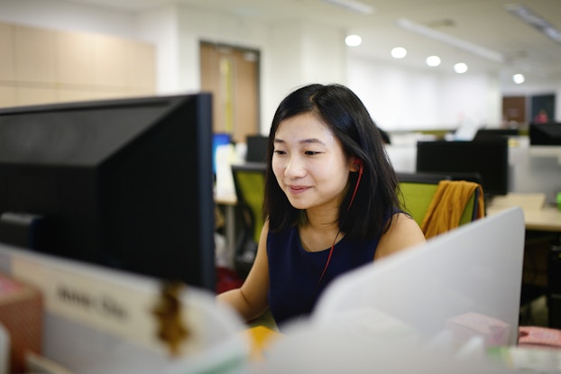  A young woman with long dark hair works at a computer in an office. She has a red wired earbud in one ear and is smiling slightly as she looks at the computer monitor. Behind her can be seen rows of desks with similar computer monitors.