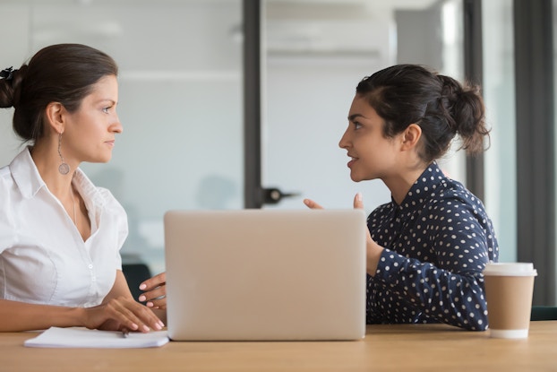 Two women sitting at a table are seen in profile, as they are turned to face each other. The woman on the left wears a white button-up blouse and has her hair in a bun. The woman on the right wears a dark blue blouse with white polka-dots. She also has her hair in a bun and she is talking and gesticulating. A laptop sits on the table between the two women, and a coffee cup sits next to the woman on the right. Behind the women is a wall made of frosted glass.
