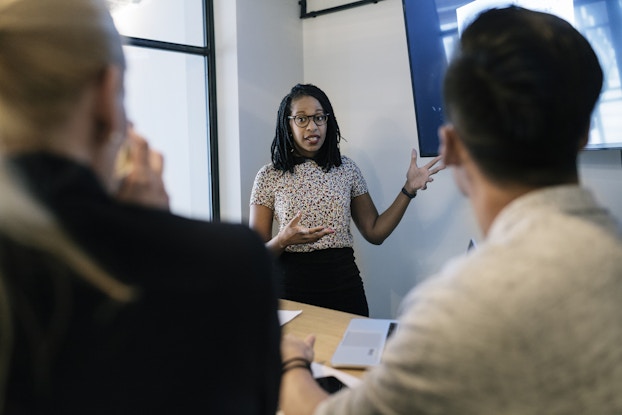 A young woman stands at the front of a conference room, speaking and gesturing with her hands. In the foreground are two people seated at the conference room table; they are seen out-of-focus and from behind. The wall-mounted screen beside the standing woman is partially obscured by the head of one of the seated people.