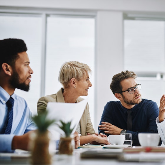 A group of five people sit around a conference table. Everyone's attention is on the fourth person from the left, a woman who is excitedly talking and gesturing with her hands.